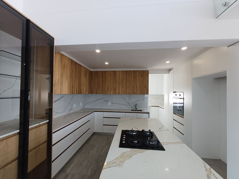 Modern kitchen featuring sleek white and wood-grain cabinetry, marble-look stone countertops, a central island with a gas cooktop, and integrated oven.