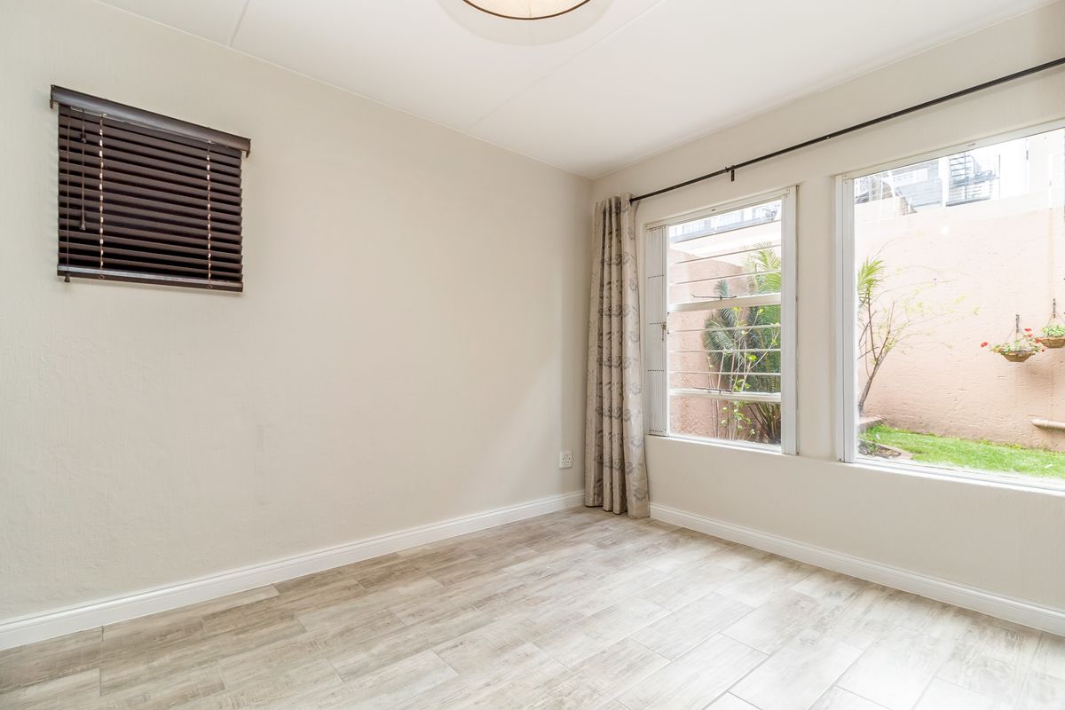 A bright, empty bedroom featuring light wood-look tile flooring, neutral walls, and large windows overlooking a private rear courtyard area.