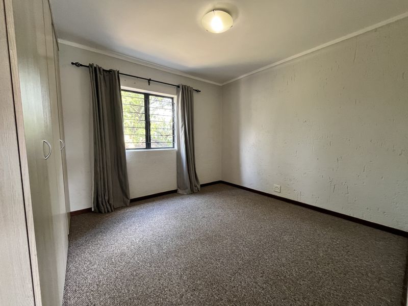 A simple, empty bedroom featuring built-in cupboards, carpeted flooring, and a window with security bars.