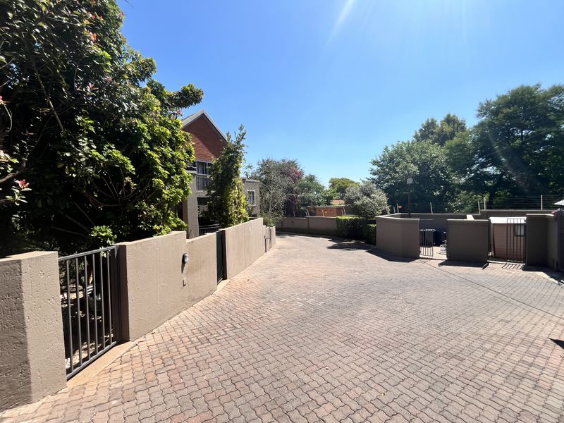 A paved driveway leading to the apartment complex entrance, featuring perimeter walls with integrated electric fencing and mature landscaping.