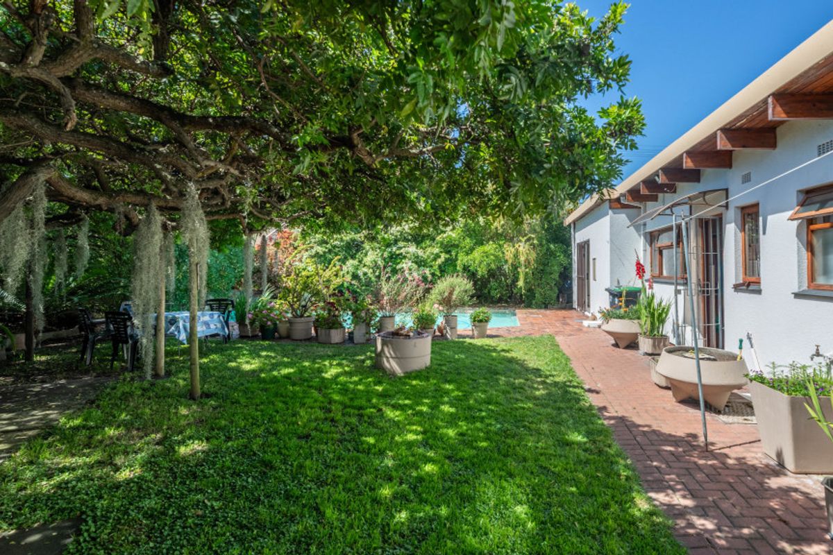 A private rear garden featuring a lush lawn, mature tree canopy, brick-paved patio area, and a swimming pool in the background.