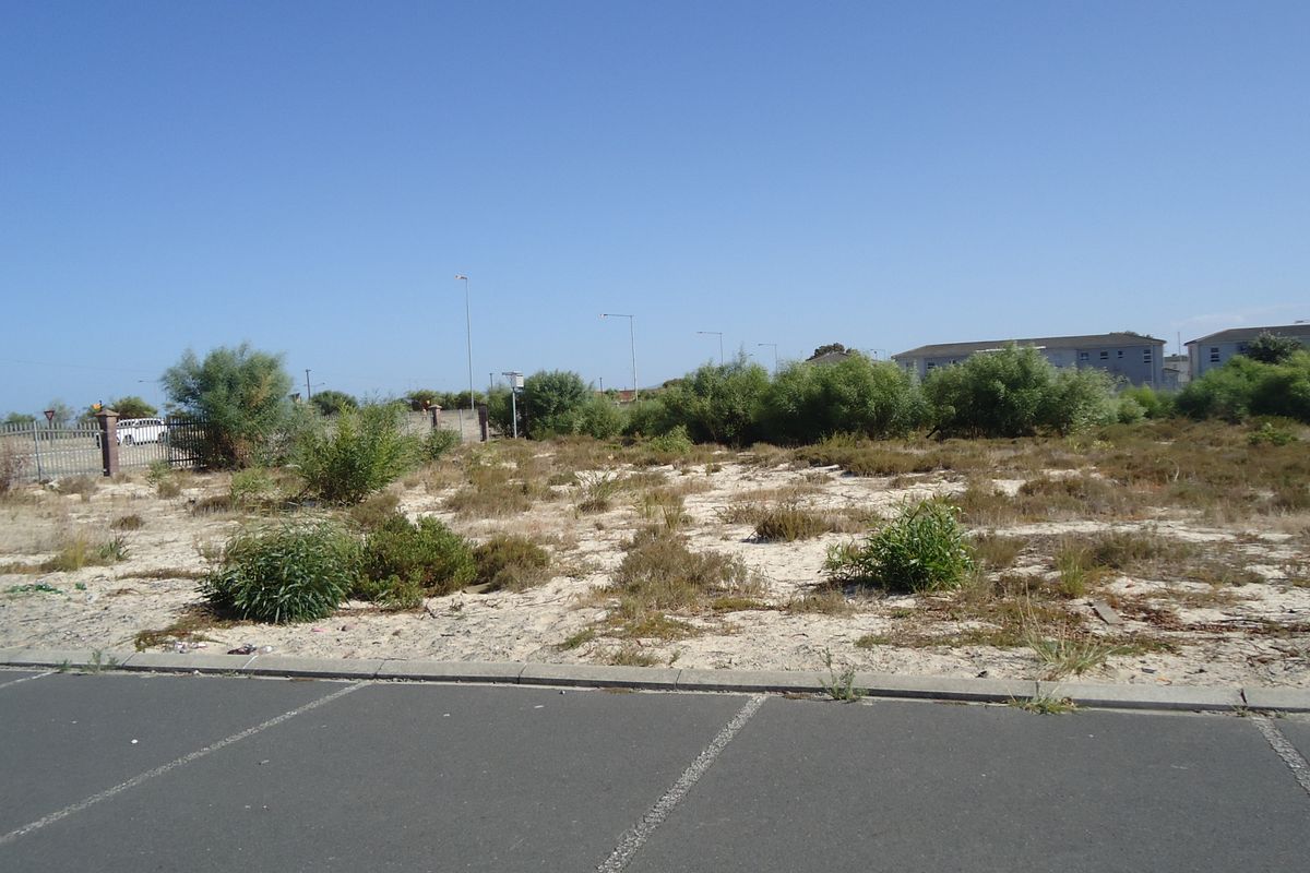A vacant residential plot of land featuring sandy terrain, natural vegetation, and perimeter fencing, situated adjacent to an asphalt road.