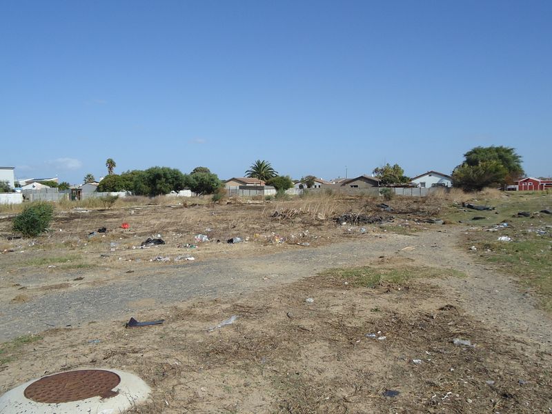 A vacant residential plot featuring a manhole cover, perimeter fencing, and surrounding neighborhood views, currently requiring site clearing and maintenance.