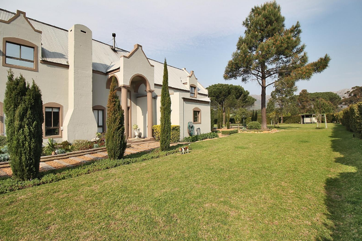 A well-maintained rear exterior view of a small holding property featuring a gabled roof, covered entrance, and expansive landscaped garden with mature trees.