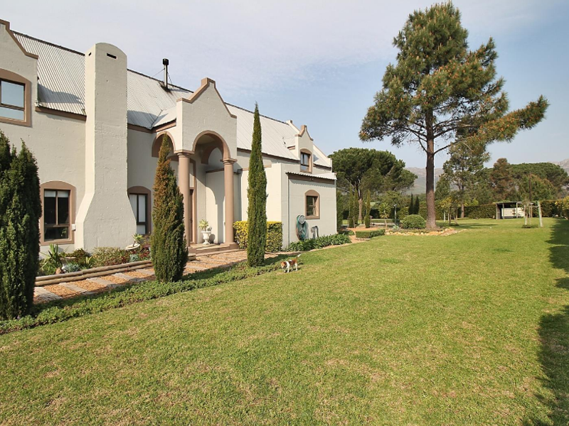 A well-maintained rear exterior view of a small holding property featuring a gabled roof, covered entrance, and expansive landscaped garden with mature trees.