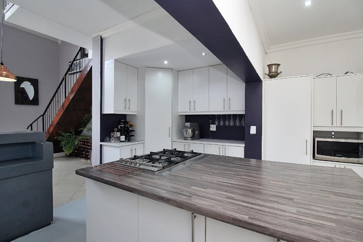Modern kitchen featuring a central island with a gas hob, ample white cabinetry, and a sleek laminate countertop, adjacent to an open staircase.