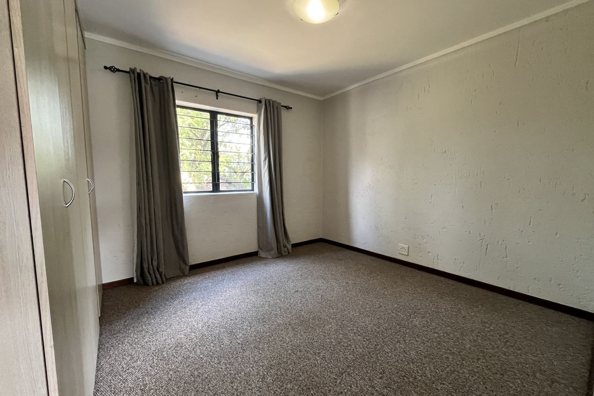A simple, empty bedroom featuring built-in cupboards, carpeted flooring, and a window with security bars.