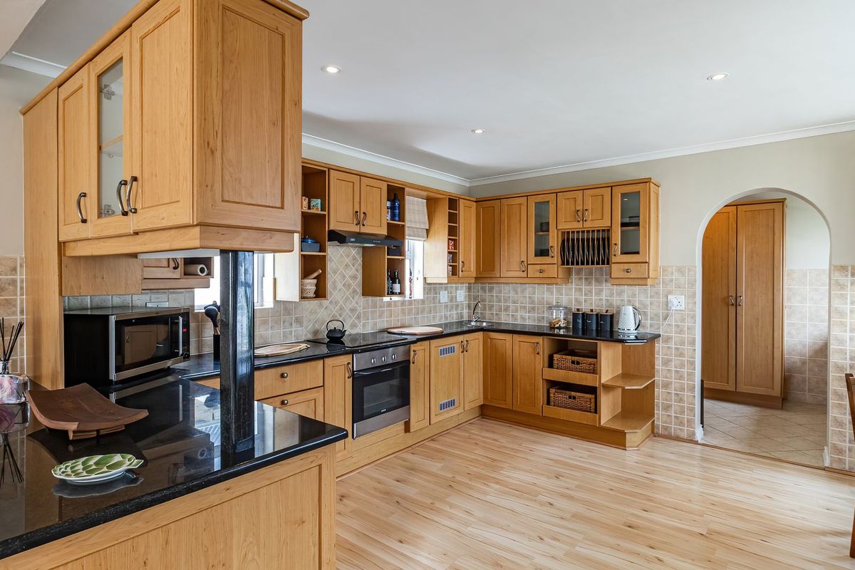 A functional kitchen featuring light wood cabinetry, dark granite countertops, tiled backsplash, and integrated oven and hob, with an arched doorway leading to adjacent space.