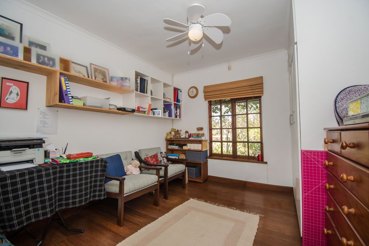 A functional bedroom or home office featuring timber flooring, built-in shelving, a ceiling fan, and a window with a timber frame.