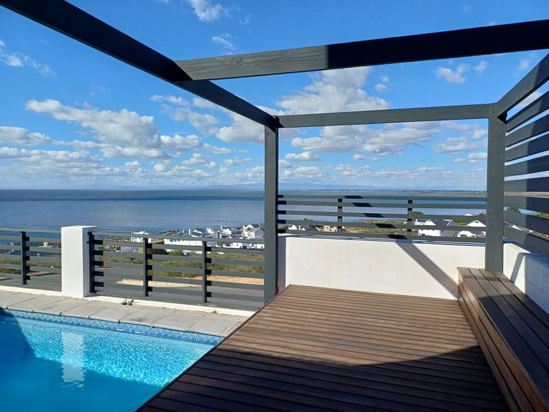 An elevated outdoor pool area featuring timber decking, a built-in bench, and a pergola, offering expansive coastal and neighbourhood views.