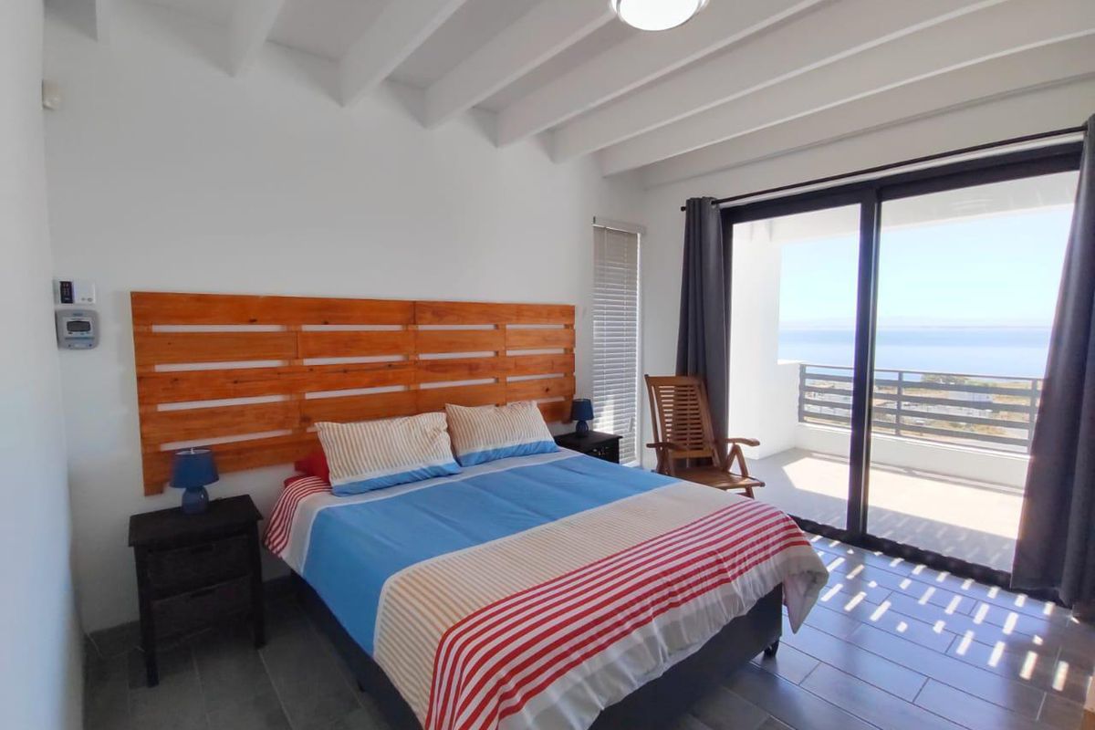 A bright bedroom featuring exposed beam ceilings, tiled flooring, and large sliding glass doors that open onto a balcony with ocean views.