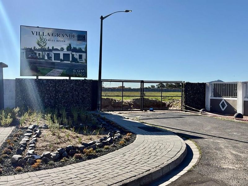 Front entrance view of the Villa Grande development site featuring a paved driveway, security gate, and gabion wall boundary.