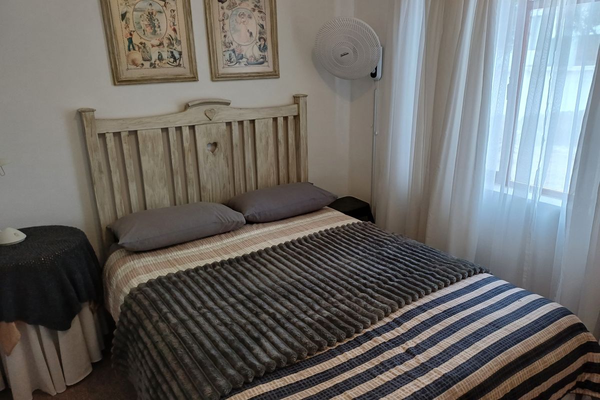 A cozy bedroom featuring a wooden headboard, wall-mounted fan, and large window with sheer curtains, providing natural light.