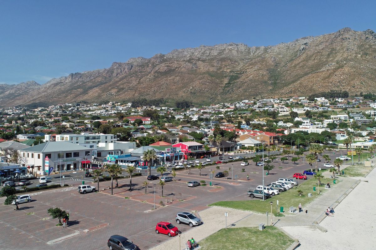 Aerial view of a coastal town featuring a large paved parking area, surrounding commercial buildings, and a scenic mountain range backdrop.
