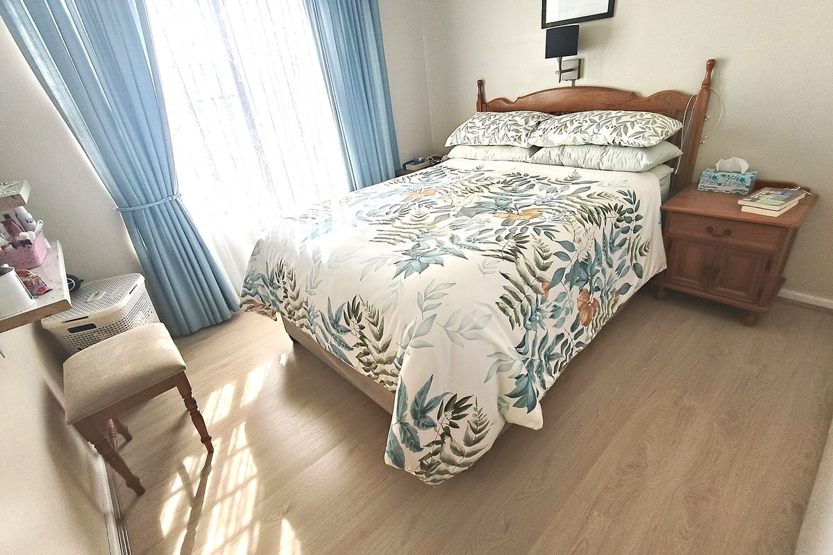 A well-lit bedroom featuring light-toned laminate flooring and large windows with blue curtains, providing a comfortable and functional sleeping space.