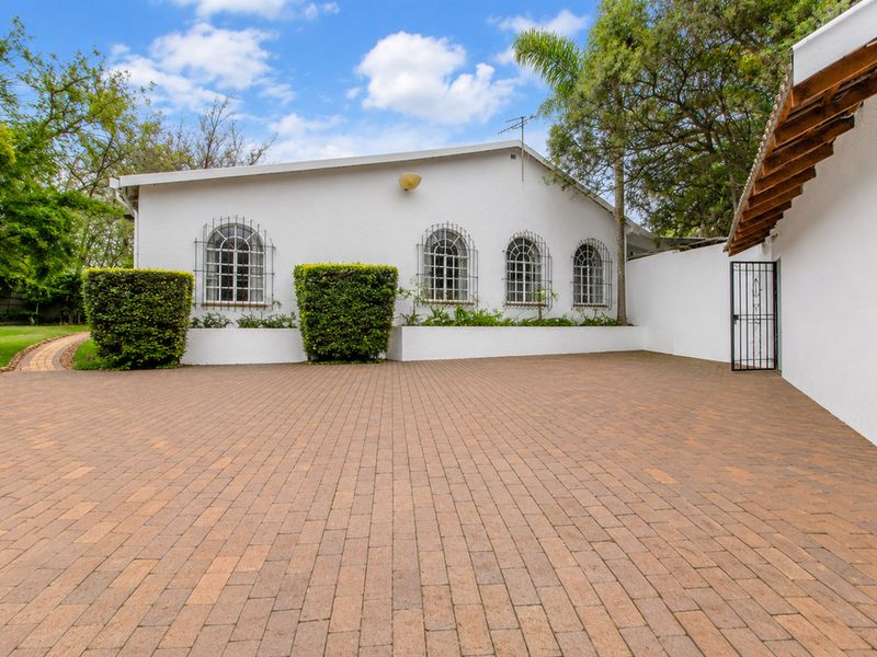 A paved driveway area leading to the front facade of the house, featuring arched windows with security grilles and established garden landscaping.