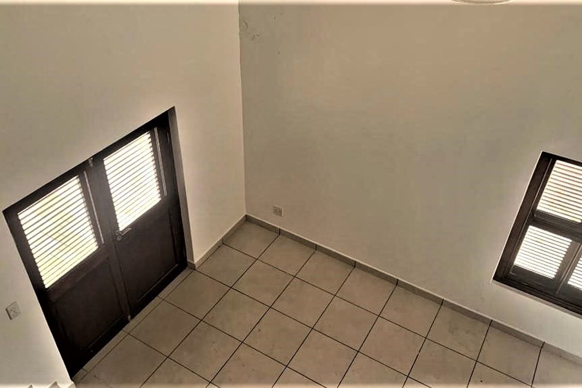 An empty, high-ceilinged room featuring tiled flooring, dark-framed double doors, and a window with integrated shutters.