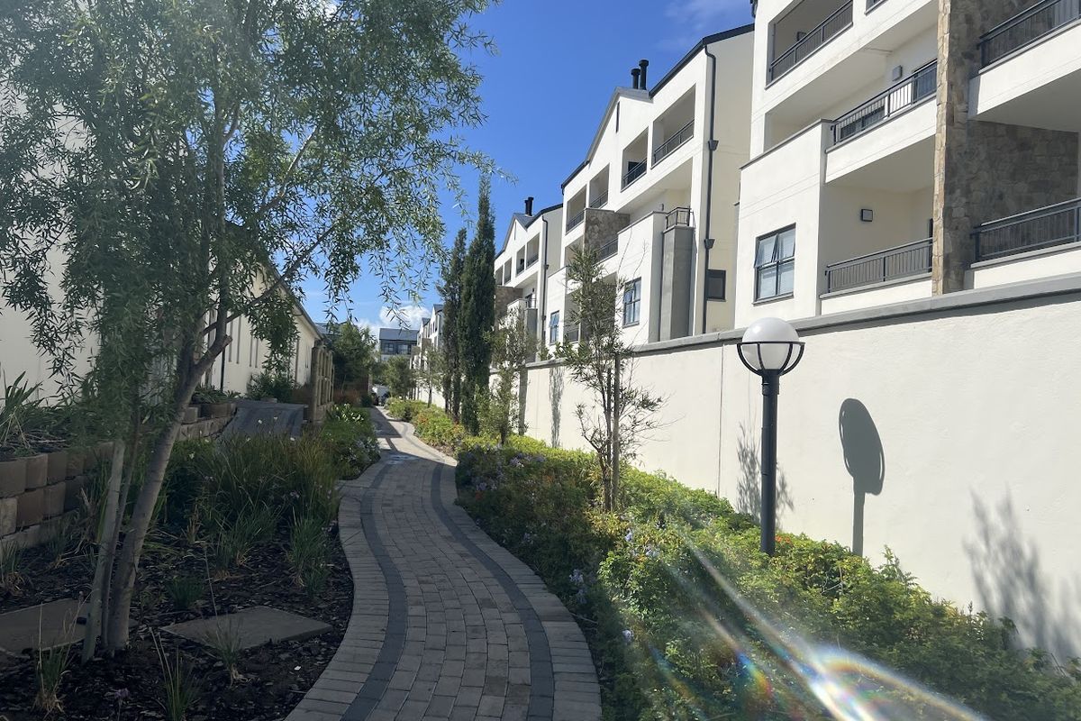 A well-maintained paved walkway winds through a landscaped garden area alongside the rear elevation of a modern apartment complex.