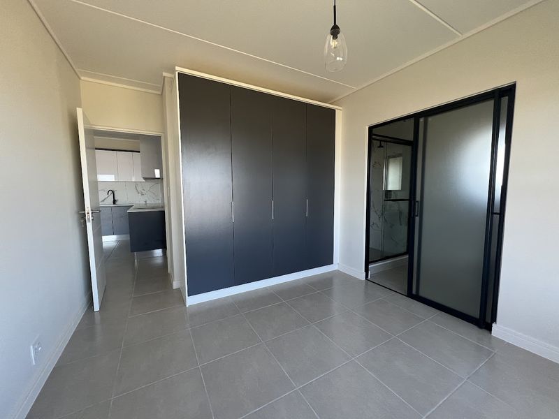 Modern bedroom featuring neutral tiled flooring, dark built-in cupboards, and a sleek sliding glass door leading to an en-suite bathroom.