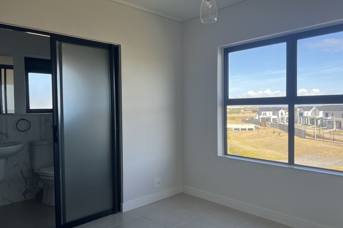A bright, modern bedroom featuring neutral tiled flooring, a large window with neighbourhood views, and a sleek sliding frosted glass door leading to an en-suite.