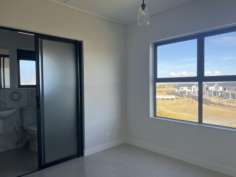 A bright, modern bedroom featuring neutral tiled flooring, a large window with neighbourhood views, and a sleek sliding frosted glass door leading to an en-suite.