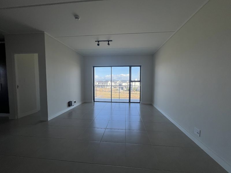 A bright, empty living room featuring neutral tiled flooring, modern track lighting, and a large sliding glass door leading to an outdoor balcony.