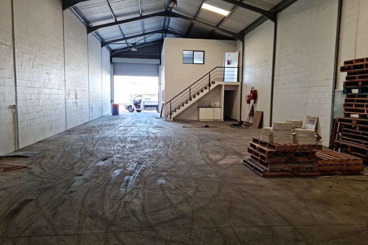 Spacious commercial warehouse unit featuring high ceilings, exposed steel trusses, a mezzanine level with staircase, and a large roller shutter door for easy access.