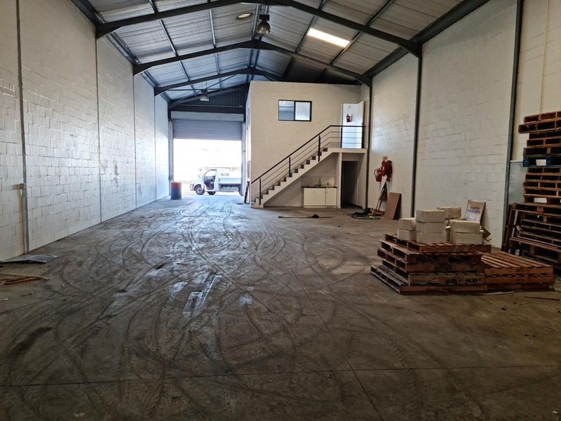 Spacious commercial warehouse unit featuring high ceilings, exposed steel trusses, a mezzanine level with staircase, and a large roller shutter door for easy access.