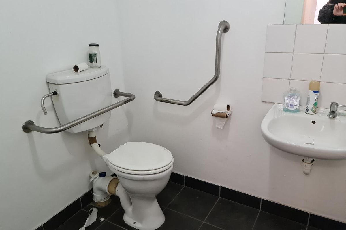Functional commercial bathroom featuring a toilet, wall-mounted basin, and stainless steel grab rails for accessibility, finished with dark floor tiles.