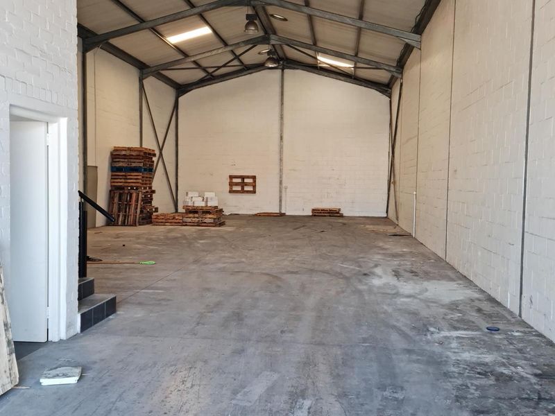 Spacious industrial warehouse or utility space featuring high ceilings with exposed steel trusses, concrete flooring, and durable painted brick walls.