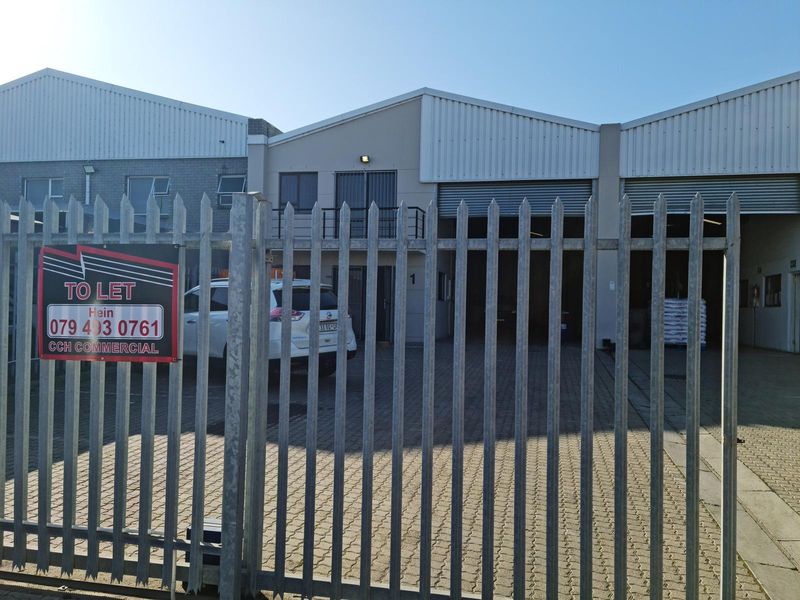 Front elevation of a commercial warehouse unit featuring roller shutter doors, paved driveway access, and secure steel palisade fencing.