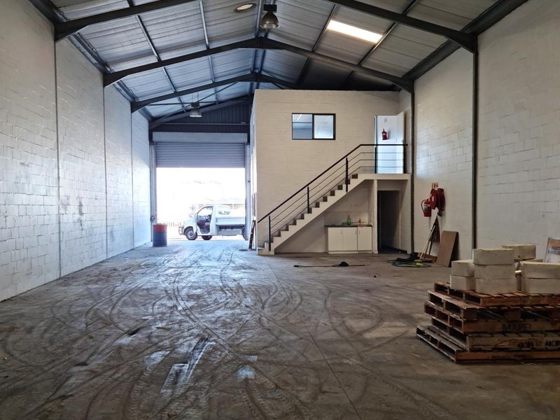 Spacious commercial warehouse featuring high ceilings with exposed steel trusses, a mezzanine level with staircase, and a large roller shutter door for vehicle access.
