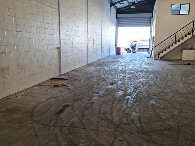 Spacious commercial warehouse or utility space featuring high ceilings with exposed steel trusses, concrete flooring, and a large roller shutter door for vehicle access.