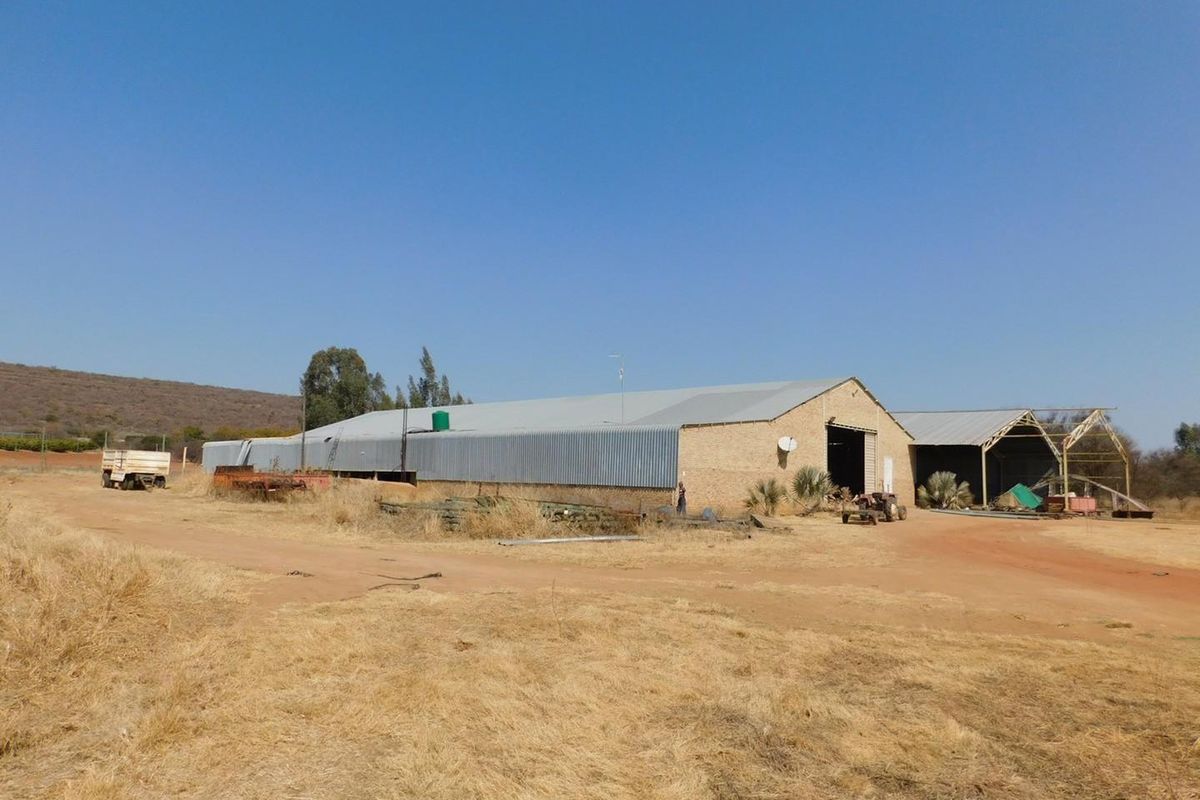 A large agricultural farm building with corrugated metal roofing and brick construction, featuring an adjacent open-sided storage structure on a rural property.