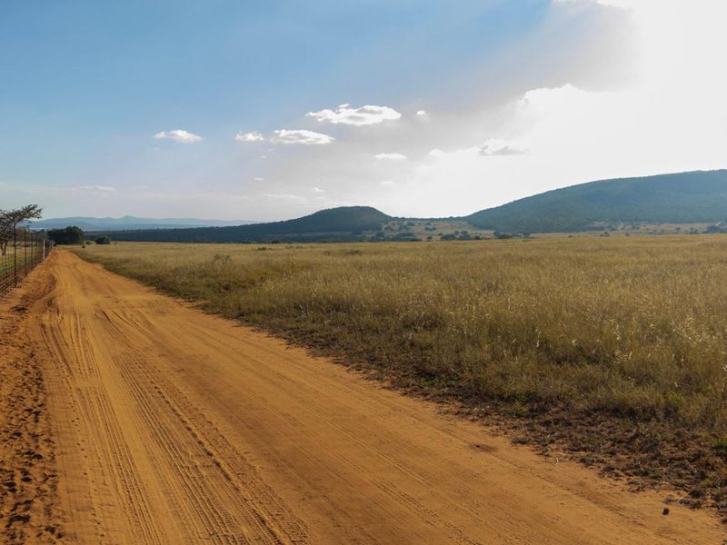 A wide-angle view of a vacant commercial land plot featuring a dirt access road and perimeter wire fencing against a scenic mountain backdrop.