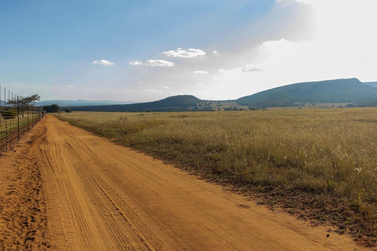 A wide-angle view of a vacant commercial land plot featuring a dirt access road and perimeter wire fencing against a scenic mountain backdrop.