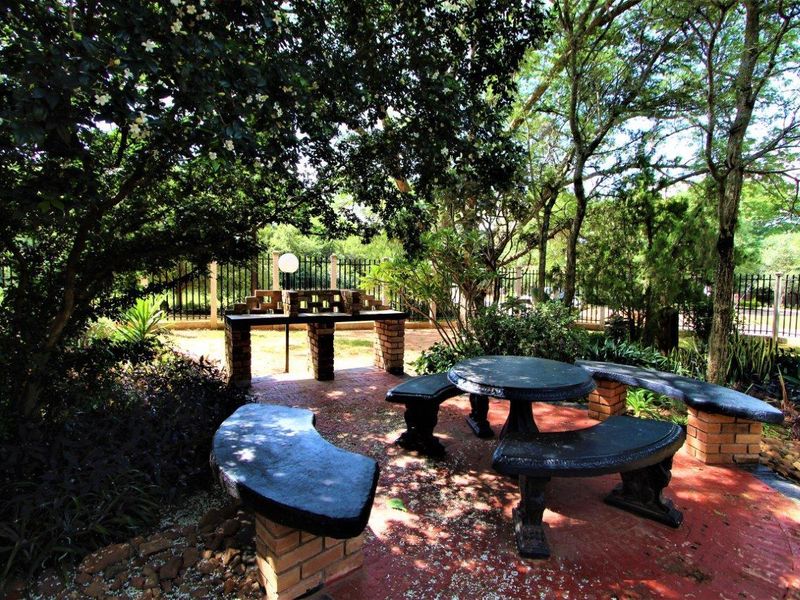A landscaped garden area featuring a brick built-in braai, paved patio space, and permanent stone bench seating under mature trees.
