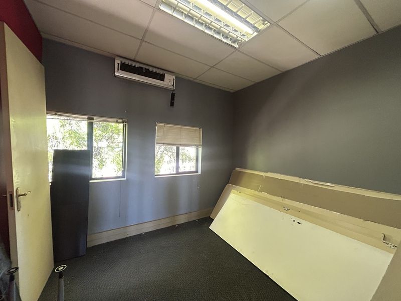 A vacant office space featuring grey walls, carpeted flooring, a suspended ceiling with integrated lighting, and a wall-mounted air conditioning unit.