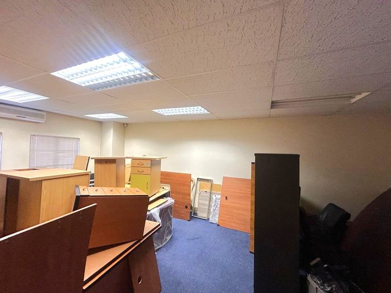 An office space featuring a suspended ceiling with recessed lighting, wall-mounted air conditioning, and carpeted flooring, currently used for storage.