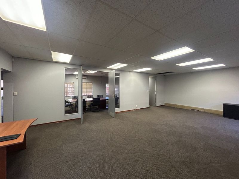 Spacious open-plan office area featuring a suspended ceiling with recessed lighting, carpeted flooring, and glass-paneled doors leading to an adjacent meeting room.