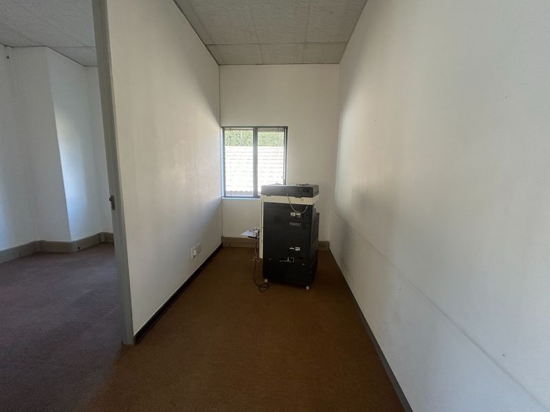 A small, empty office space featuring carpeted flooring, a suspended ceiling, and a window with blinds, suitable for a private workspace.