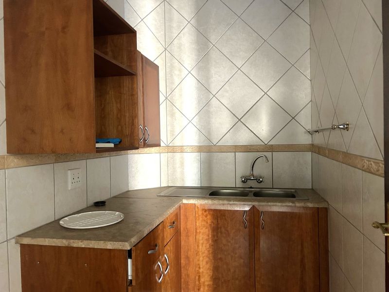 A compact office kitchenette featuring wooden cabinetry, laminate countertops, and a stainless steel double sink with tiled wall finishes.