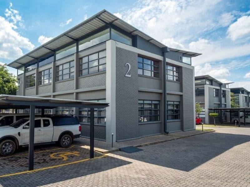 Modern multi-story office building featuring a grey brick facade, clerestory windows, and dedicated covered parking bays with paved driveway access.