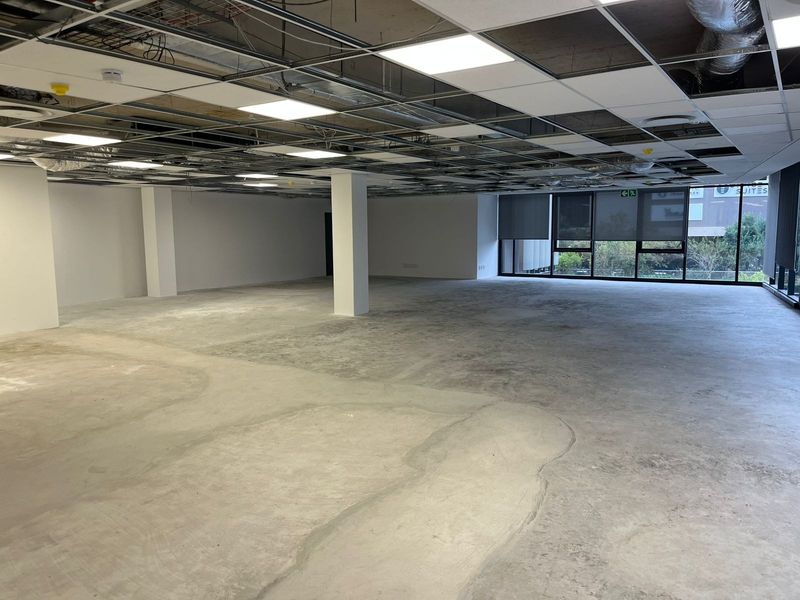 An expansive, open-plan office space currently in a shell condition, featuring exposed ceiling infrastructure, concrete flooring, and large windows providing natural light.