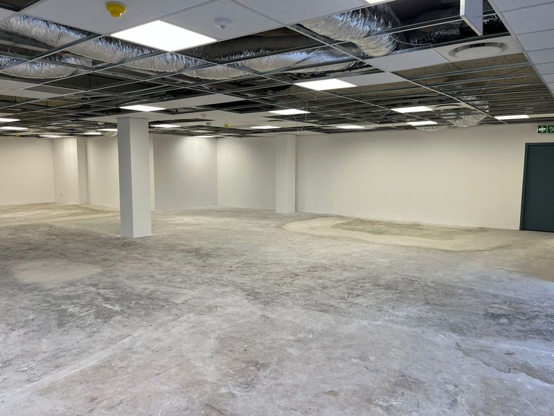 An open-plan office space currently under renovation, featuring an exposed ceiling grid, structural columns, and unfinished concrete flooring.
