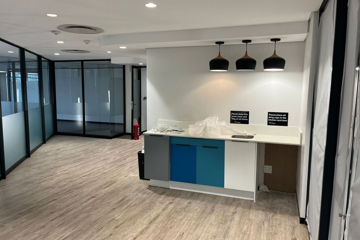 A modern office kitchenette area featuring laminate flooring, glass partitions, pendant lighting, and built-in cabinetry with a sink.
