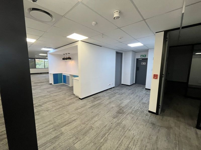 A modern, open-plan office space featuring a built-in kitchenette, suspended ceilings with recessed lighting, and neutral carpeted flooring.