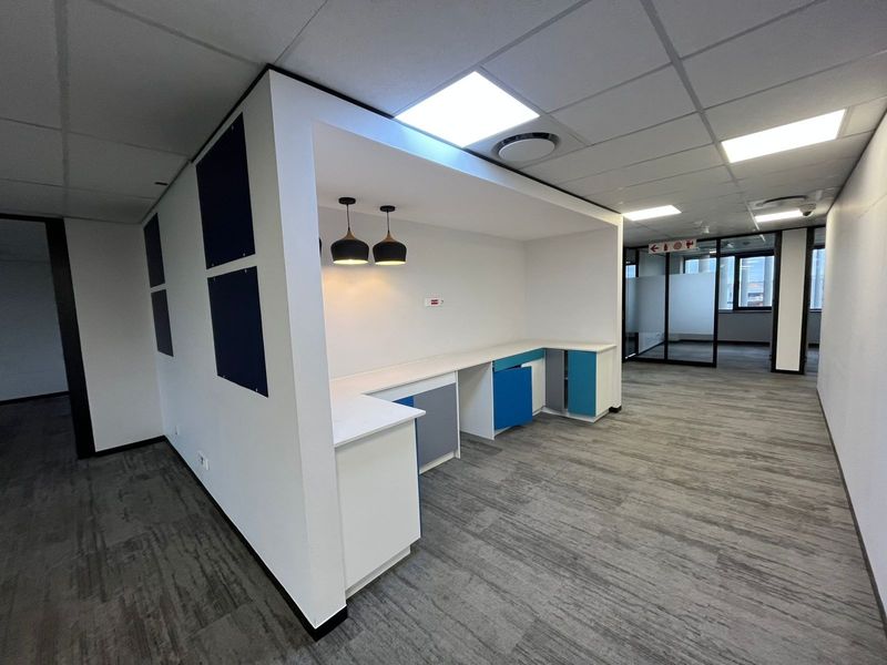Modern open-plan office space featuring built-in kitchenette cabinetry, suspended ceilings with recessed lighting, and durable carpet tiles, offering a clean and professional work environment.