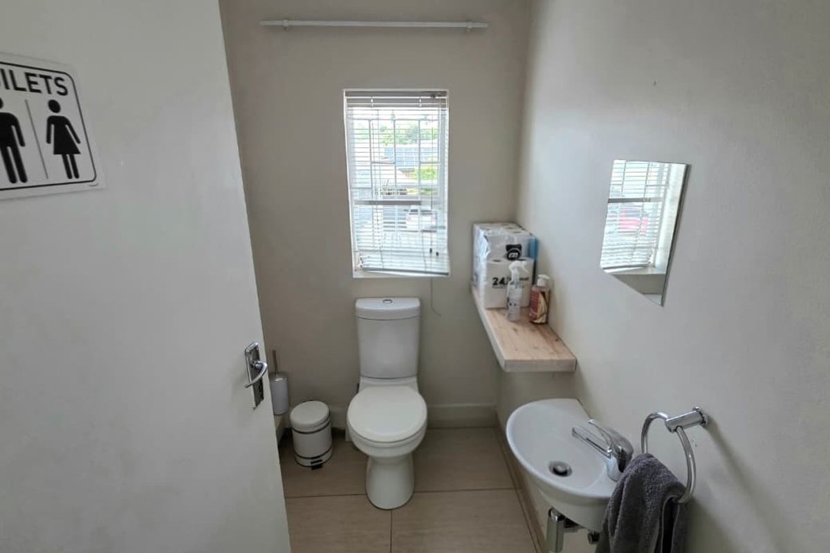 A functional office restroom featuring a toilet, wall-mounted basin, chrome fixtures, and tiled flooring with natural light from a window.