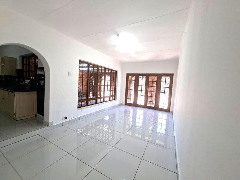 A bright, open-plan office space featuring polished white tiled flooring, an arched entryway, and large wooden-framed windows and doors providing ample natural light.